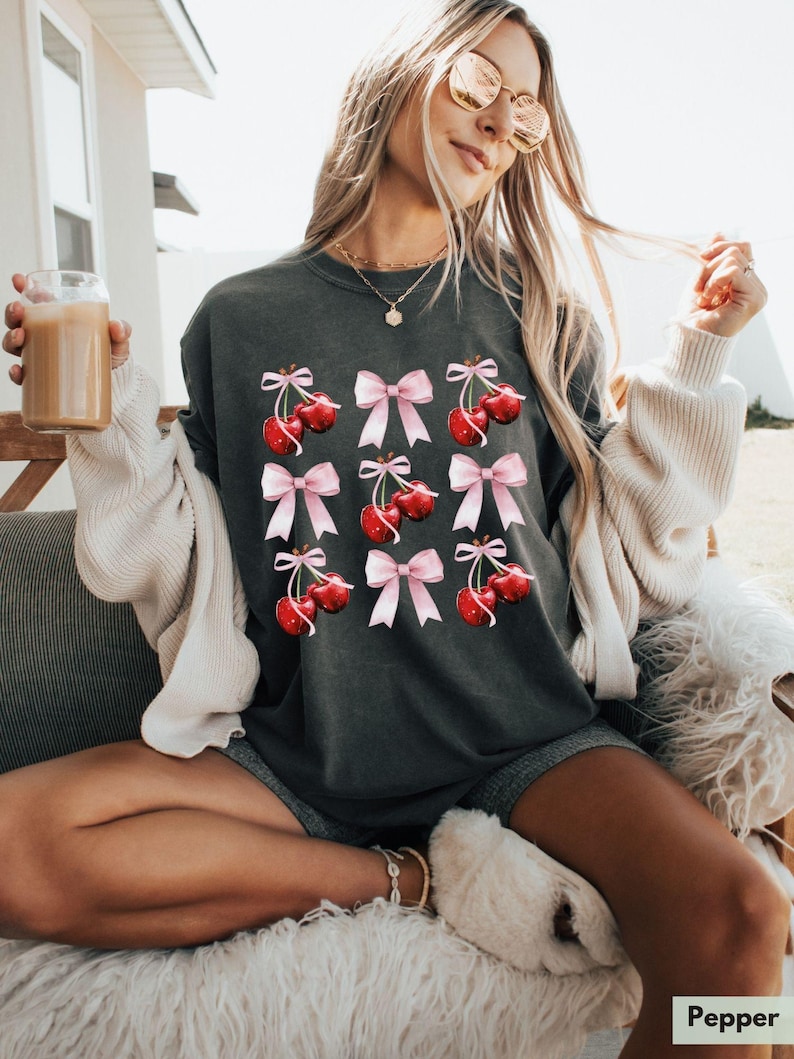 May include: A dark gray t-shirt with a graphic of red cherries with pink bows. The cherries are arranged in a grid pattern.