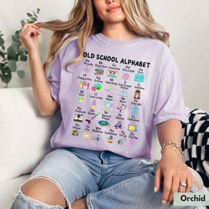 May include: A purple t-shirt with the text "OLD SCHOOL ALPHABET" and a colorful alphabet chart featuring retro items like a boombox, a hula hoop, a record player, and a walkman.