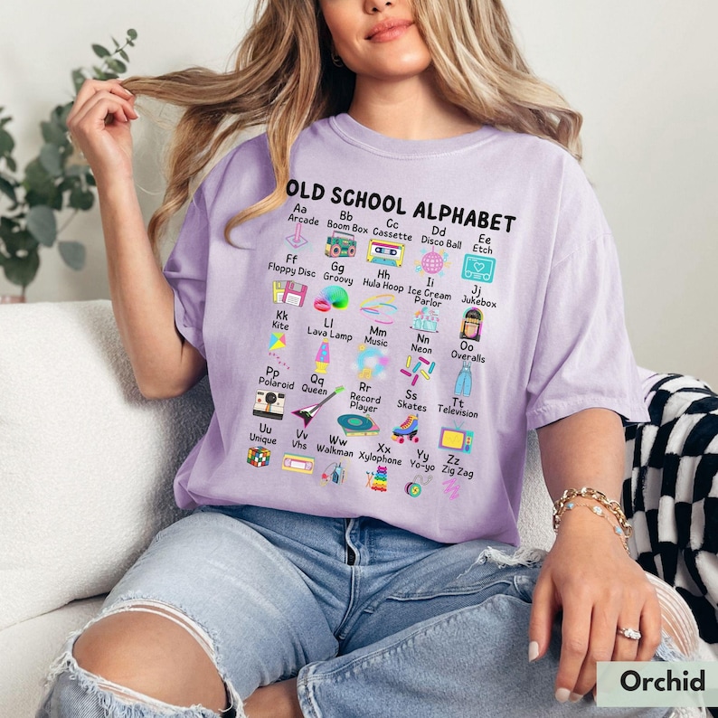 May include: A purple t-shirt with the text "OLD SCHOOL ALPHABET" and a colorful alphabet chart featuring retro items like a boombox, a hula hoop, a record player, and a walkman.