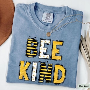 May include: A blue jean colored t-shirt with the words "BEE KIND" in yellow and black block letters. The letter "E" in "BEE" is filled with a sunflower pattern. A gold necklace with a small pendant rests on the shirt.