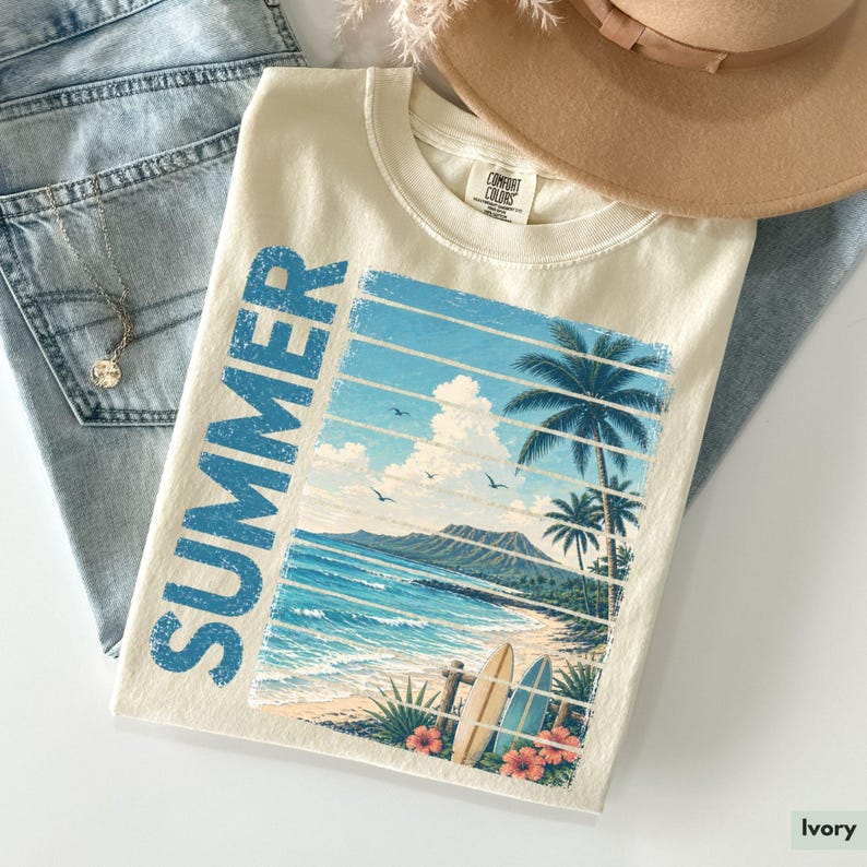 May include: Ivory-colored t-shirt with a beach scene graphic. The design features a blue ocean, palm trees, and surfboards, with the word "SUMMER" in blue. A tan hat and blue jeans are also in the image.