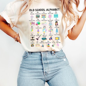 May include: A white t-shirt with a graphic of an alphabet printed on it. The graphic features illustrations of objects from the 1970s and 1980s, such as a boombox, cassette tape, hula hoop, and arcade game. The text "OLD SCHOOL ALPHABET" is printed at the top of the graphic.