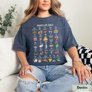 May include: A woman wearing a blue t-shirt with a white graphic of farm-related items and the text "FARM LIFE ABCS". The graphic includes illustrations and letters representing different farm-related words, such as "Agriculture", "Barn", "Chickens", "Dairy", "Eggs", "Fence", "Grain", "Hay", "Irrigation", "Jam", "Kettle", "Lamb", "Milk", "Nest", "Organic", "Plow", "Quail", "Rooster", "Scarecrow", "Tractor", "Udder", "Vegetables", "Wheelbarrow", "Xyris", "Yield", and "Zucchini".