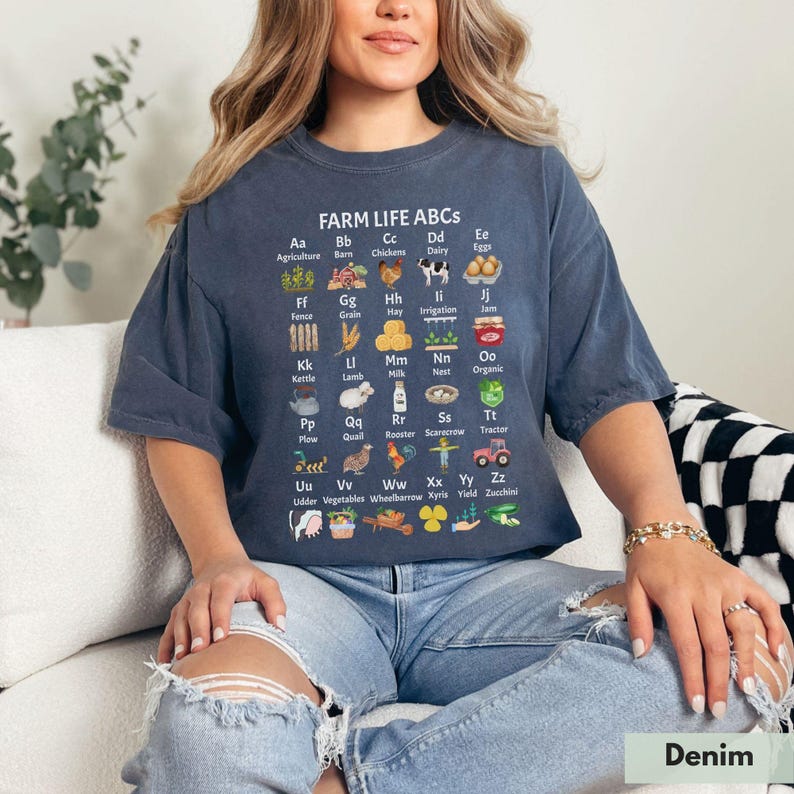 May include: A woman wearing a blue t-shirt with a white graphic of farm-related items and the text "FARM LIFE ABCS". The graphic includes illustrations and letters representing different farm-related words, such as "Agriculture", "Barn", "Chickens", "Dairy", "Eggs", "Fence", "Grain", "Hay", "Irrigation", "Jam", "Kettle", "Lamb", "Milk", "Nest", "Organic", "Plow", "Quail", "Rooster", "Scarecrow", "Tractor", "Udder", "Vegetables", "Wheelbarrow", "Xyris", "Yield", and "Zucchini".