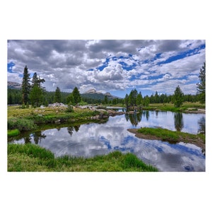 Puede incluir: Un paisaje sereno con un lago tranquilo que refleja el cielo azul y nubes blancas y esponjosas. Árboles verdes bordean la orilla, y un pico de montaña se eleva en la distancia.