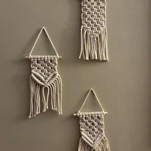 May include: Three white macrame wall hangings with fringe, each with a different pattern. The hangings are attached to wooden dowels.