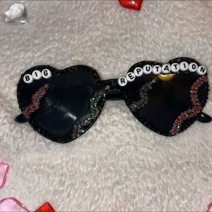 Puede incluir: Gafas de sol negras en forma de corazón con detalles de purpurina rosa y plateada. Las gafas tienen las palabras "BIG" y "REPUTATION" escritas en cuentas blancas.