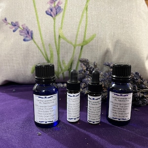 May include: Four blue glass bottles of essential oil with black caps, labeled "Carolina Lavender Farm." A linen bag with a lavender sprig design is in the background, along with a bundle of dried lavender. The bottles contain 0.5 fl oz and 1 fl dram of oil.