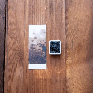 May include: A small square white plastic pan filled with dark charcoal-coloured watercolour paint, accompanied by a rectangular white paper swatch showing a gradient of the same dark pigment, ranging from a light wash to a dense, opaque application.