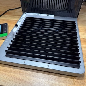 May include: A silver and black honeycomb laser bed with a white corner stop guide labeled XTOOL. The device features a grid of black metal slats and is connected to a small rectangular control panel with a screen displaying a tree icon.