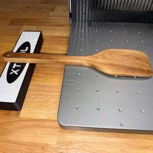 May include: A light brown wooden spatula with a flat, wide head and a long handle. The spatula is positioned horizontally across a grey metal base plate featuring a grid of small circular holes and a white rectangular block labeled xTool.