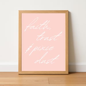 Pode incluir: Uma impressão emoldurada com fundo rosa claro e texto branco em itálico que diz "faith, trust, & pixie dust". A moldura é de madeira clara. A impressão é exibida em um piso de madeira.