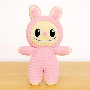 Puede incluir: Un juguete de peluche de ganchillo rosa y amarillo hecho a mano con forma de monstruo con orejas de conejo. El juguete tiene ojos negros grandes, una boca sonriente con dientes blancos y brazos y piernas pequeños. El peluche está hecho de hilo suave y texturizado.