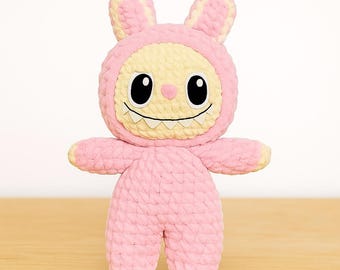 May include: A handmade pink and yellow crochet monster toy with bunny ears. The toy features large black eyes, a smiling mouth with white teeth, and small arms and legs. The plush is made of soft, textured yarn, perfect for children.