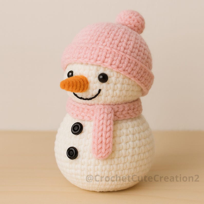 Puede incluir: Un mu&ntilde;eco de nieve tejido a ganchillo con gorro y bufanda rosas, una nariz de zanahoria y ojos de bot&oacute;n negros. El mu&ntilde;eco de nieve tiene una boca sonriente y est&aacute; hecho a mano. El texto "@CrochetCuteCreation2" est&aacute; en la parte inferior.