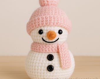 May include: A crocheted snowman figure with a pink hat and scarf, black button eyes, and a carrot nose. The snowman is white with pink accents and a smiling face. The text "@CrochetCuteCreation2" is at the bottom. A cute winter decoration.