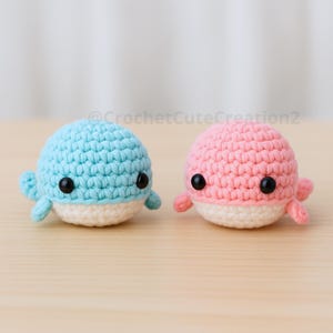 Puede incluir: Dos figuras de ballenas de ganchillo. Una es azul claro y la otra rosa. Ambas tienen ojos negros y vientres blancos. Las ballenas son pequeñas, hechas a mano y están sobre una superficie marrón claro.