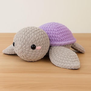 Puede incluir: Una tortuga de peluche hecha a mano con cabeza y extremidades grises, caparazón lavanda y ojos negros. La tortuga tiene un rubor rosa claro en las mejillas y está hecha de hilo suave y texturizado. Este adorable peluche es perfecto para niños.