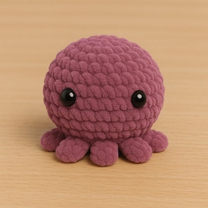 Puede incluir: Un juguete de pulpo de peluche hecho a mano en un tono púrpura apagado. El pulpo presenta una cabeza redonda con dos ojos grandes de botón negro y ocho tentáculos cortos y redondeados. El juguete está hecho de un material tejido texturizado.