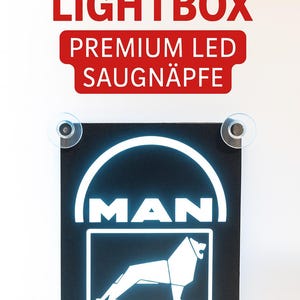 Könnte beinhalten: Schwarze LED-Leuchtbox mit MAN-Logo und Löwen-Grafik. Der Schriftzug "MAN TRUCKS LIGHTBOX" ist rot, ebenso wie "PREMIUM LED SAUGNÄPFE". Das Schild wird von vier Saugnäpfen gehalten.