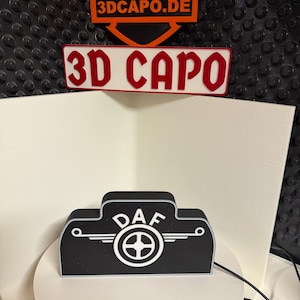 May include: Black and white 3D printed DAF logo with a white steering wheel and wings. The logo is on a white pedestal with a black background.