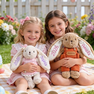 Può includere: Due bambine sorridenti, ognuna con un coniglietto di peluche. Un coniglietto è bianco con un vestito rosa e il nome "Sophia" sull'orecchio, l'altro è marrone con una tuta arancione e il nome "Amelia" sull'orecchio. Entrambi i coniglietti hanno "2026" ricamato sulle orecchie. Tema pasquale.