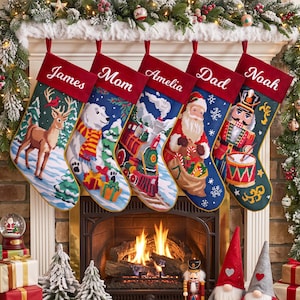 Op de afbeelding: Vijf gepersonaliseerde kerstsokken hangen aan een schoorsteenmantel. Elke sok heeft een dieprode boord met een wit geborduurde naam: James, Mom, Amelia, Dad en Noah. De ontwerpen bevatten een hert, een ijsbeer, een trein, de Kerstman en een notenkraker.