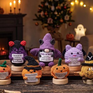 May include: A collection of seven crocheted Halloween-themed figures on wooden bases. The figures include pumpkins, a bat, a vampire, and a gnome, each holding a small white sign with printed text. The items feature yarn in orange, purple, black, and light purple.