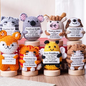 Pode incluir: Uma coleção de figuras de animais de crochê feitas à mão, incluindo um gato, um coala, uma vaca, uma preguiça, uma raposa, um tigre, uma abelha e outro gato. Cada figura segura uma placa com afirmações positivas.