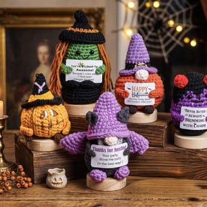 May include: Five crocheted Halloween figures on wooden stands. One is a green witch, one an orange pumpkin, one a purple-hatted gnome, one a purple bat-winged gnome, and one a purple vampire. Each holds a sign with text including "Wicked-ly Awesome", "HAPPY Halloween", "NEVER BITES", and "Thanks for hosting".