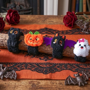 Op de afbeelding: Vier pluche polsbandjes met Halloween-figuren: een zwarte kat, een oranje pompoen, een zwarte vleermuis met paarse vleugels en een wit spookje met een roze strik. Elk figuurtje is bevestigd aan een zwarte, pluizige band op een houten stam.