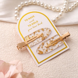 May include: Two gold-coloured hair clips with clear, oval resin centres containing gold leaf flakes. They are displayed on a white and gold card with the text "TO HAVE and TO HOLD your hair back". A pearl necklace is in the background.