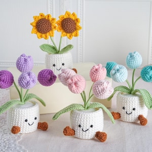 Puede incluir: Una colección de flores de ganchillo hechas a mano en macetas blancas con caras sonrientes. Las flores incluyen girasoles, tulipanes morados, rosas y azules. Las macetas tienen pies marrones y hojas verdes.