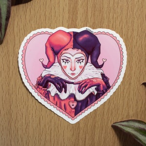 May include: A sticker of a jester with a red and black jester hat, wearing a white ruff, and holding their hands together in front of their chest. The jester is inside a pink heart with a white outline and a scalloped edge. The sticker has the text "NISAVAC" in the top right corner.