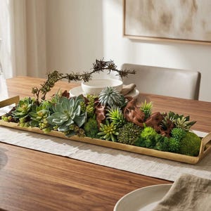 May include: A long, rectangular gold-toned metal tray with handles at each end, filled with an arrangement of various artificial green succulents, moss, and pieces of brown driftwood, creating a dense, textured botanical centerpiece.