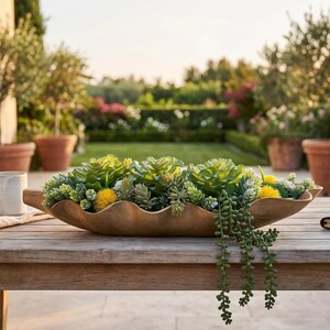 May include: A long, wavy-edged wooden bowl filled with an assortment of artificial green succulents and small yellow flowers. The bowl sits on a wooden table next to a white ceramic mug, a folded napkin, and a closed notebook with a pen on top.