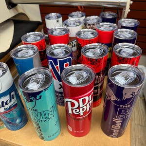 May include: A collection of insulated tumblers with various designs and logos. The tumblers feature brands like Red Bull, Dr. Pepper, and others. They are cylindrical with clear lids and are displayed on a wooden surface.
