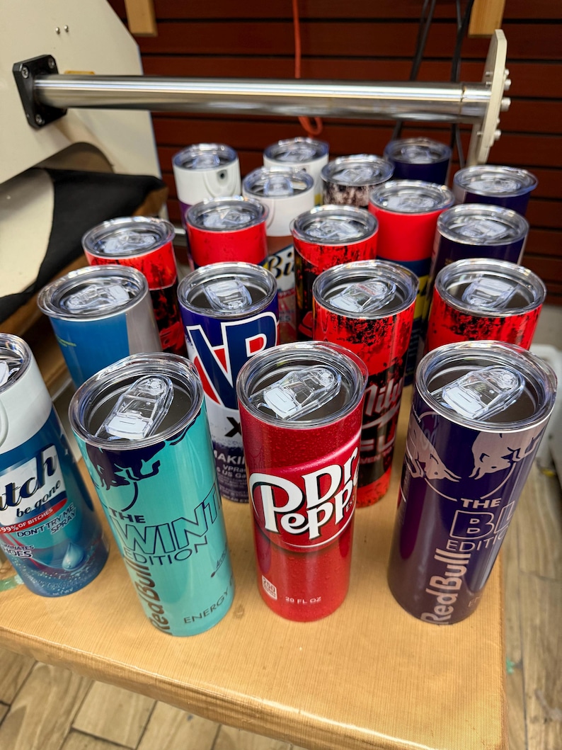 May include: A collection of insulated tumblers with various designs and logos. The tumblers feature brands like Red Bull, Dr. Pepper, and others. They are cylindrical with clear lids and are displayed on a wooden surface.