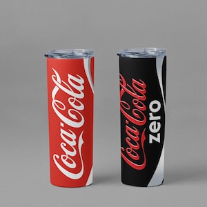 May include: Two insulated tumblers, one red with white "Coca-Cola" script and the other black with white "Coca-Cola Zero" script. Both have clear lids and a silver accent. The tumblers are cylindrical and likely hold approximately 20 ounces.