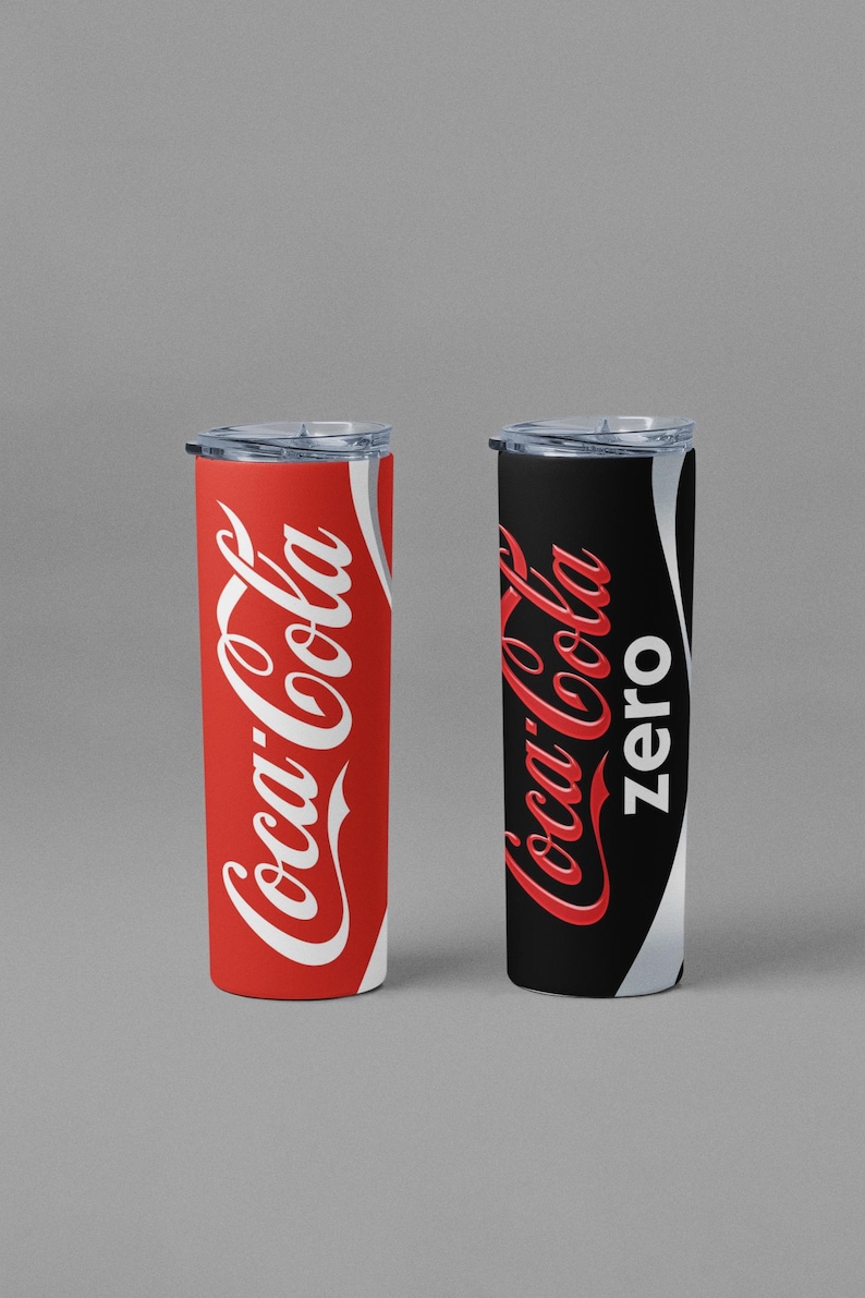 May include: Two insulated tumblers, one red with white "Coca-Cola" script and the other black with white "Coca-Cola Zero" script. Both have clear lids and a silver accent. The tumblers are cylindrical and likely hold approximately 20 ounces.