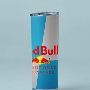 May include: A cylindrical, insulated tumbler with a clear lid. The tumbler features a blue and gray color scheme with the Red Bull logo and text. The text reads "ENERGY DRINK SUGARFREE" and "VITALIZES BODY AND MIND".