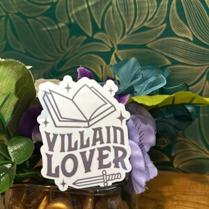 Puede incluir: Una pegatina con un libro y una espada, y el texto "VILLAIN LOVER" en fuente morada.