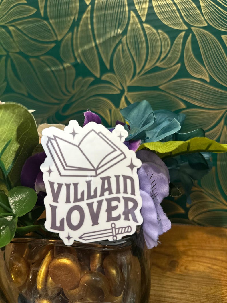 Puede incluir: Una pegatina con un libro y una espada, y el texto "VILLAIN LOVER" en fuente morada.