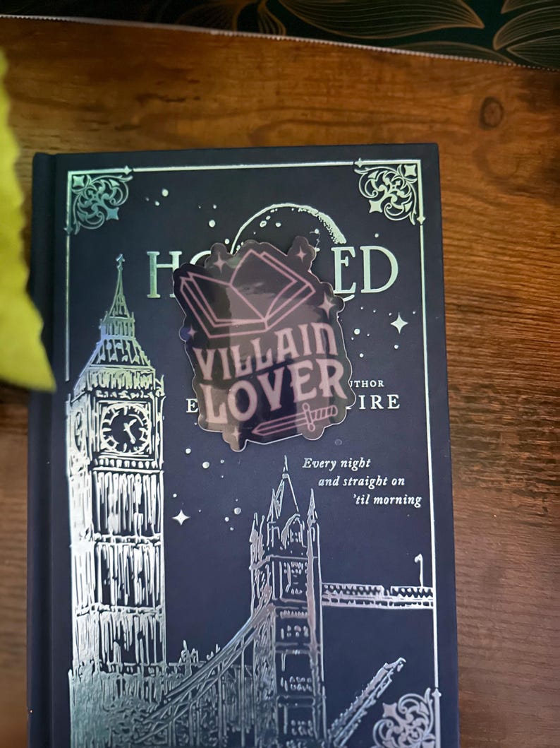Puede incluir: Una pegatina hologr&aacute;fica con un dise&ntilde;o de libro y el texto "VILLAIN LOVER" en una cubierta de libro azul oscuro con un dise&ntilde;o en relieve plateado del Tower Bridge y el Big Ben. La cubierta del libro tambi&eacute;n tiene el texto "Every night and straight on 'til morning".