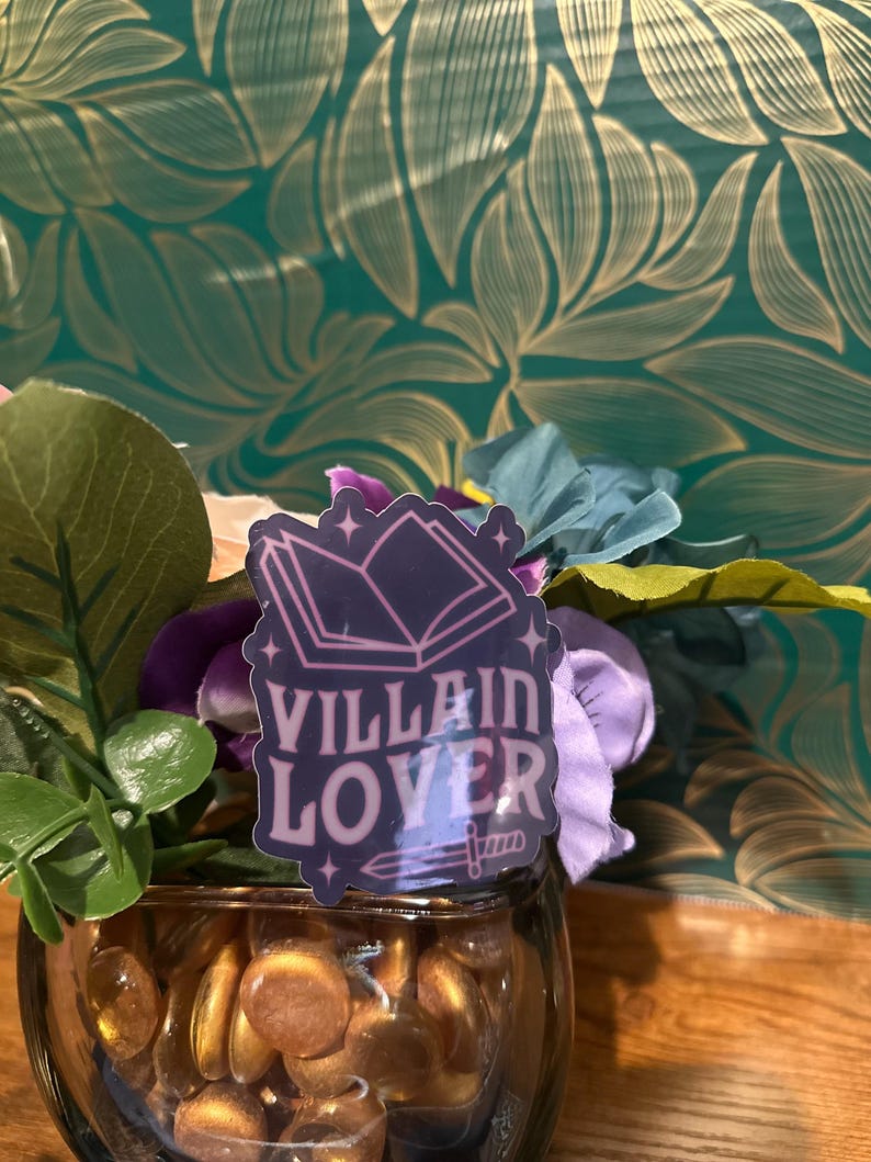 Puede incluir: Una pegatina rosa y morada con un libro abierto, una espada y el texto "Villain Lover". La pegatina est&aacute; en un jarr&oacute;n de vidrio lleno de piedras de vidrio doradas.