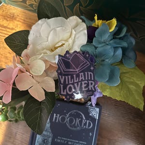 Puede incluir: Primer plano de un libro titulado "Hooked" de Emily McIntire con una pegatina que dice "Villain Lover" en la parte superior. El libro est&aacute; rodeado de un ramo de flores blancas, rosas y azules.
