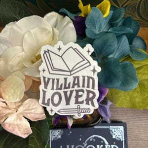 Puede incluir: Una pegatina blanca con un contorno negro de un libro abierto, estrellas y el texto "VILLAIN LOVER" con una espada debajo. La pegatina est&aacute; en un ramo de flores blancas, rosas y azules.
