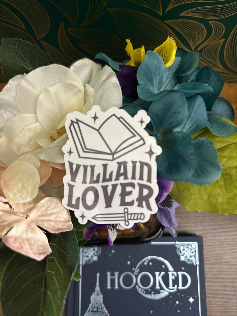 Puede incluir: Una pegatina blanca con un contorno negro de un libro abierto, estrellas y el texto "VILLAIN LOVER" con una espada debajo. La pegatina est&aacute; en un ramo de flores blancas, rosas y azules.