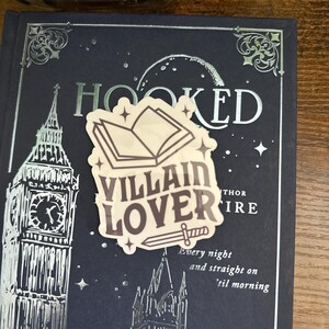 Puede incluir: Una pegatina con un libro, una espada y el texto "Villain Lover" en un libro azul oscuro con letras plateadas en relieve que dicen "Hooked".