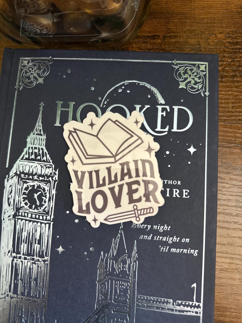 Puede incluir: Una pegatina con un libro, una espada y el texto "Villain Lover" en un libro azul oscuro con letras plateadas en relieve que dicen "Hooked".